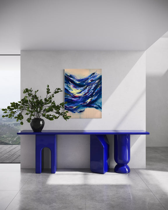An abstract canvas painting with swirling blue, navy, and white waves on a cream background, displayed on a white wall above a glossy blue console table with sculptural legs in a modern room.